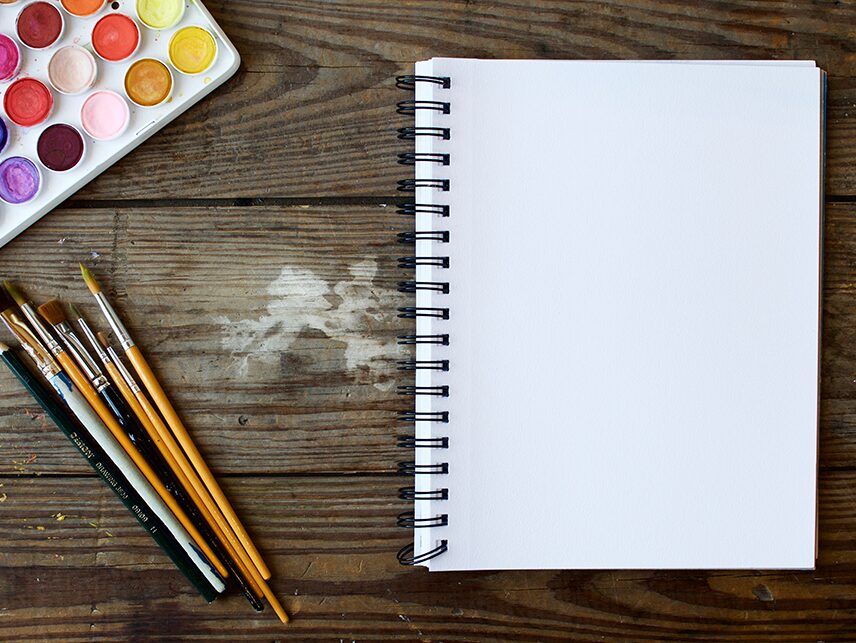 Wasserfarben, Stifte und ein Notizblock auf einem Holztisch von oben