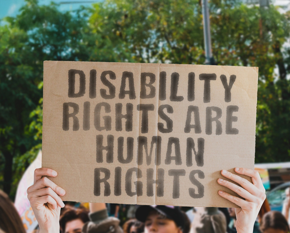 Protestplakat mit der Aufschrift „Disability Rights Are Human Rights“, gehalten von einer Menschenmenge bei einer Demonstration.