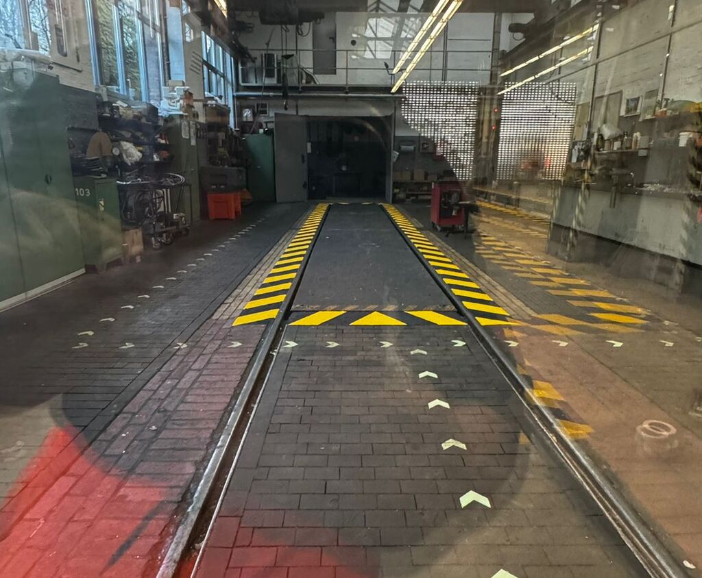 Innenaufnahme einer Werkhalle mit Schienen, gelben Sicherheitsmarkierungen und Richtungspfeilen auf dem Boden zur Orientierung und Gefahrenkennzeichnung.