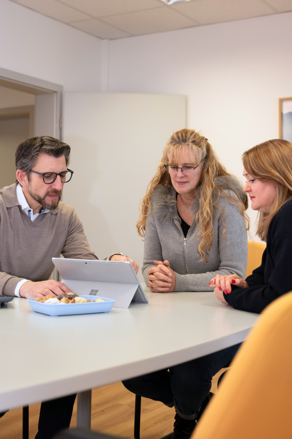 Drei Personen sitzen an einem runden Tisch, eine blonde Frau hält einen Stift, Bücher und Hefte liegen dort.