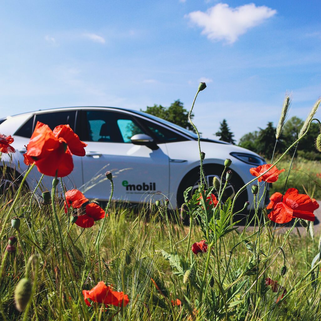 Weißes Elektroauto mit eMobil-Branding hinter roten Mohnblumen auf grüner Wiese unter blauem Himmel, Symbol für klimafreundliche Elektromobilität in der Altmark