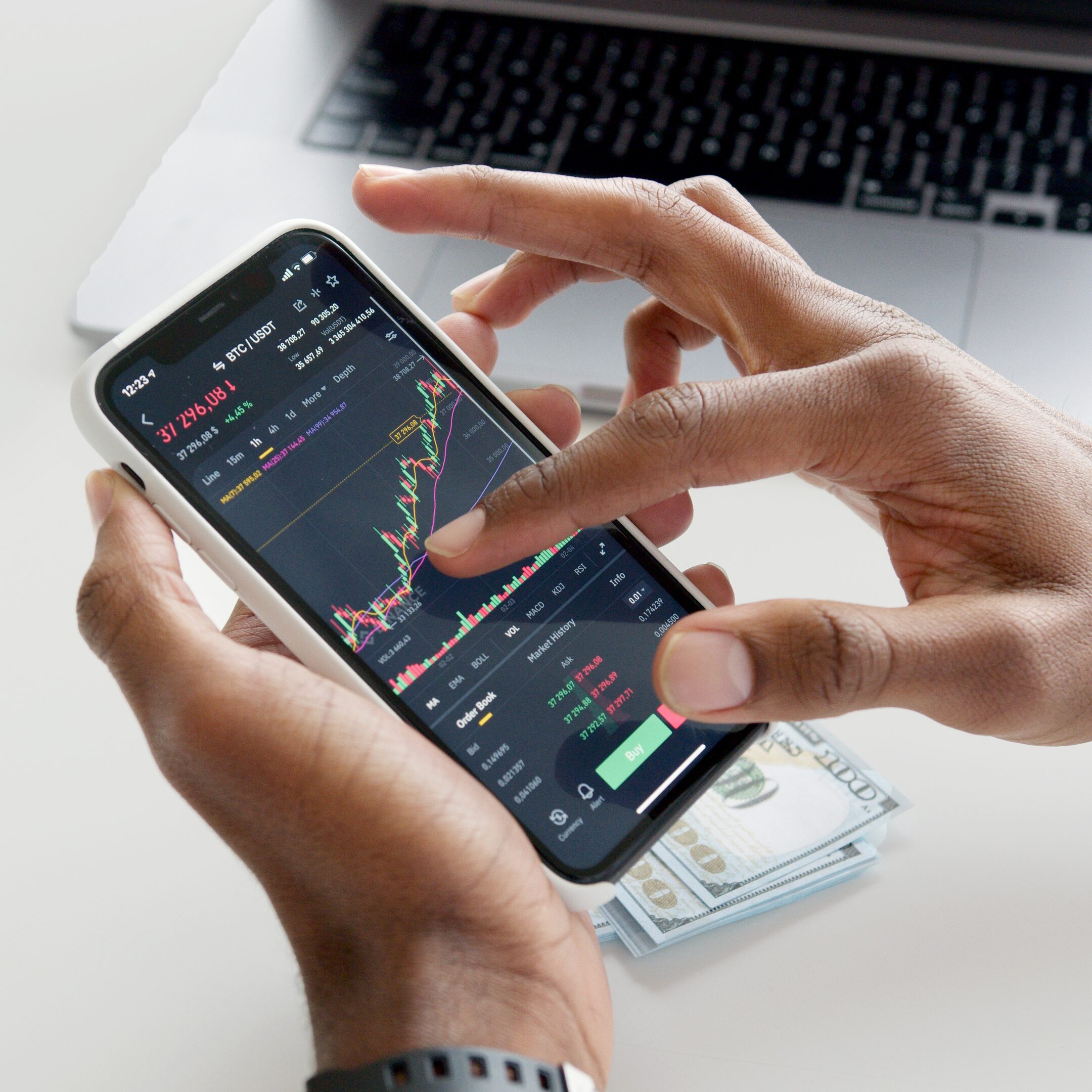 Stock image of a person of color using an investment app next to a stack of cash and a computer