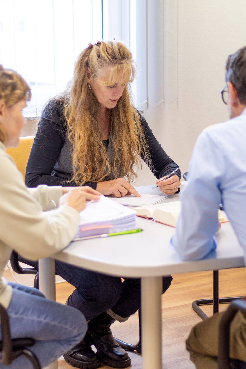 Drei Personen sitzen an einem runden Tisch, eine blonde Frau hält einen Stift, Bücher und Hefte liegen dort.
