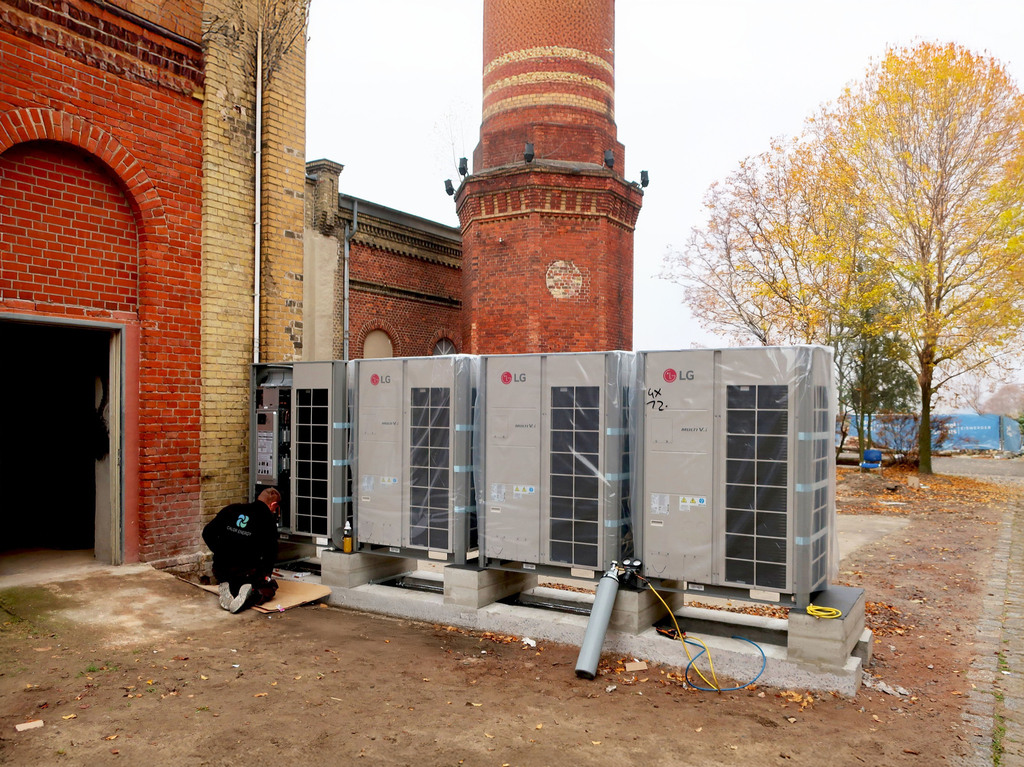 Vier Wärmepumpen-Außengeräte vor einer historischen Fabrikhalle, ein CALOR ENERGY Monteur davor
