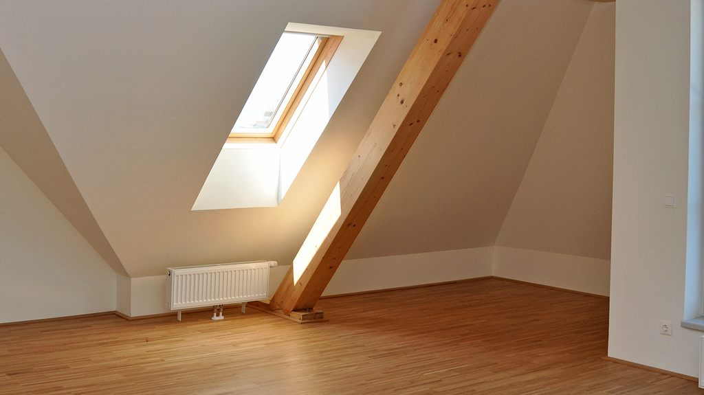 Modern ausgebautes Dachgeschoss mit Holzboden, sichtbarem Dachbalken und Dachfenster, durch das Tageslicht einfällt.