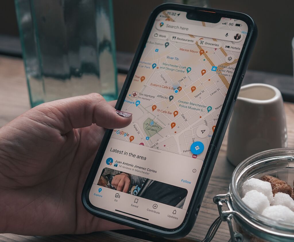Die Hand einer Frau hält ein Smartphone mit geöffneter Google Maps App. Im Hintergrund ist ein Tisch auf dem sich eine durchsichtige Vase sowie ein weißes Porzellanmilchkännchen und ein Glasbehälter mit Zuckerwürfeln darin befinden zu sehen.