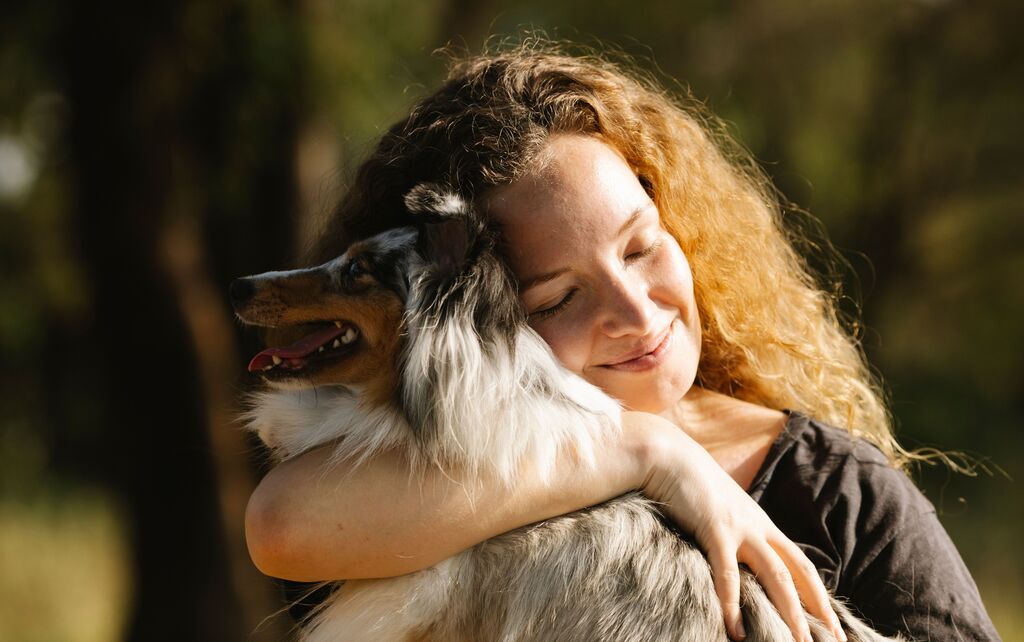 Eine rothaarige Frau umarmt lächelnd einen langhaarigen braun-weiß-grauen Hund.