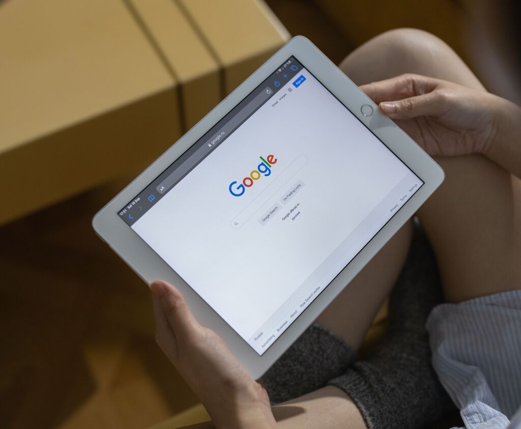 Eine Frau sitzt im Schneidersitz und hält ein weißes Tablet mit geöffneter Google-Suche-Website in den Händen.