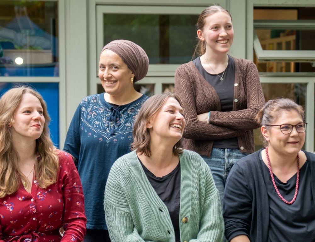 Fünf Mitarbeiterinnen stehen und sitzen lachend vor einem Gebäude