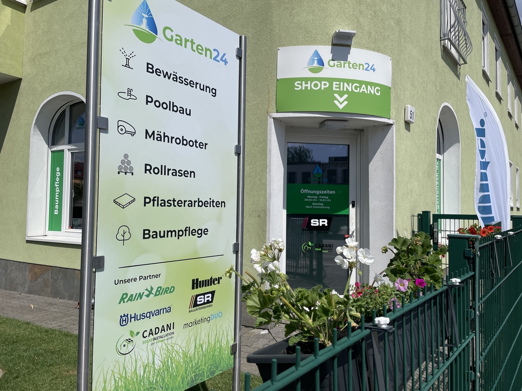 Eingang des Garten24-Shops mit einem großen Hinweisschild zu den Dienstleistungen Bewässerung, Poolbau, Mähroboter, Rollrasen, Pflasterarbeiten und Baumpflege; darunter Partnerlogos von Rain Bird, Husqvarna, Hunter, Cadani und anderen.