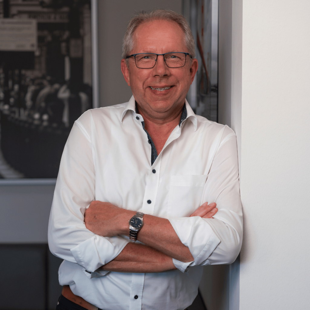 Portrait: Ernst Meyer, Mann mit Brille und grauem Haar, lächelt und steht mit verschränkten Armen in weißem Hemd vor einer Wand