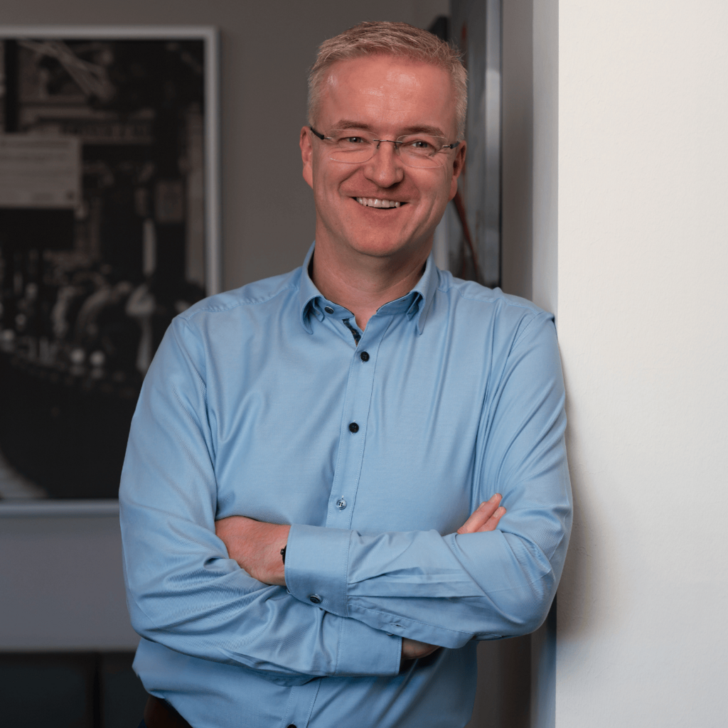 Portrait: Hendrik Meyer lehnt lächelnd an einer weißen Wand, trägt ein hellblaues Hemd und eine Brille, Arme verschränkt