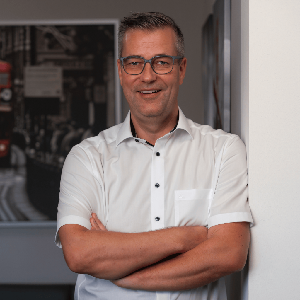 Portrait: Karsten Schmidt mit kurzen grauen Haaren, Brille und weißem Hemd, lächelnd mit verschränkten Armen an einer Wand lehnend