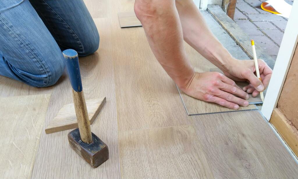 Ein Handwerker verlegt einen Laminatboden und markiert eine Schnittlinie mit einem Bleistift auf einer Holzplatte. Neben ihm liegt ein Hammer mit einem blauen Griff und ein Holzkeil, der zur Anpassung der Dielen verwendet wird. Die Szene spielt sich in der Nähe einer Türöffnung ab.