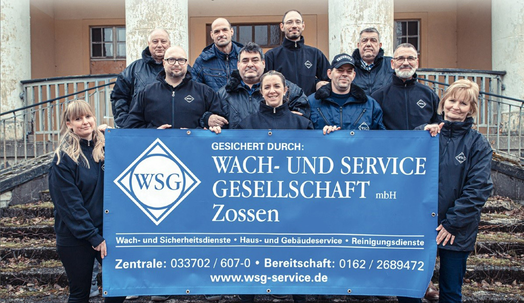 Team der WSG Zossen steht gemeinsam auf einer Treppe und hält ein großes blaues Firmenbanner mit Kontaktdaten und Leistungsbereichen.