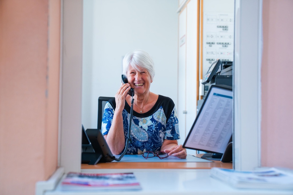 Ältere Dame mit Telefonhörer lächelt am Empfangsschalter