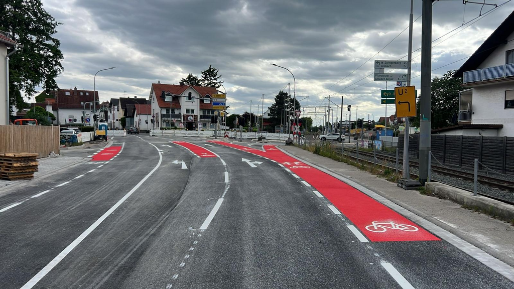 Neue Straßenmarkierungen mit weißen Fahrbahnlinien und roten Fahrradspuren in einer innerstädtischen Kreuzung, inklusive Richtungspfeilen und Radwegsymbolen.