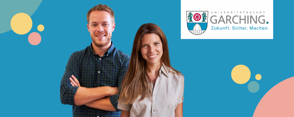 Zwei junge Fachkräfte stehen lächelnd vor dem Logo der Stadt Garching
