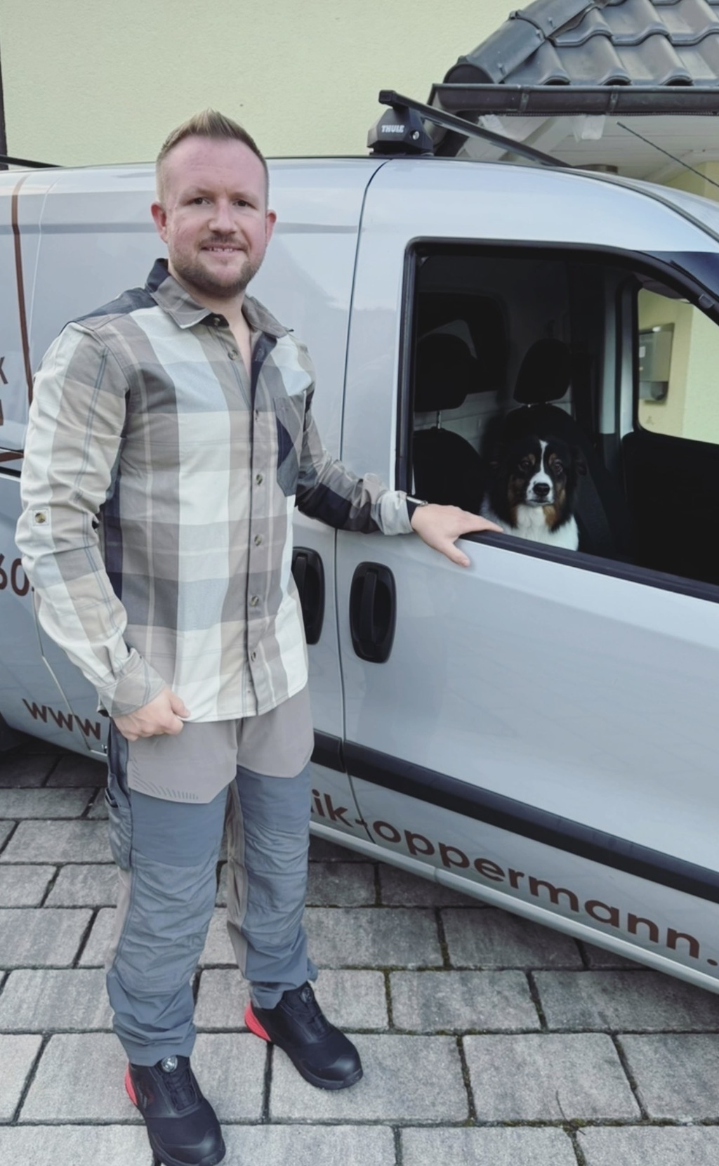 Handwerksmeister Daniel Oppermann steht vor seinem beschrifteten Firmentransporter, auf dem Beifahrersitz sitzt sein Hund