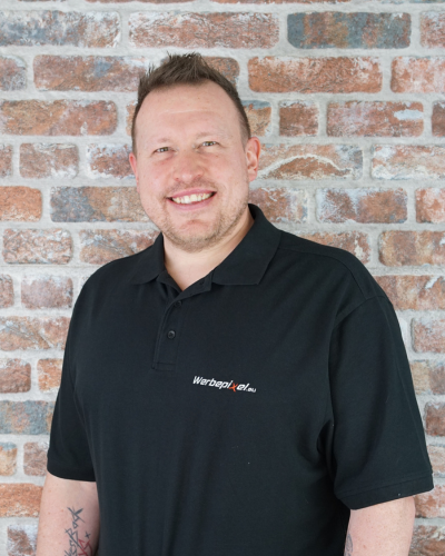 Mitarbeiterporträt von Thorsten Gilmer (Assistenz), der im Werbepixel-Poloshirt vor einer Ziegelwand lächelt