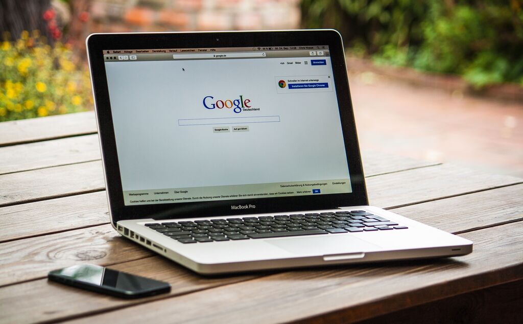 Ein silber-schwarzer Laptop auf dem die Google-Suche-Website zu sehen ist, steht auf einem Gartentisch mit einem Handy daneben.