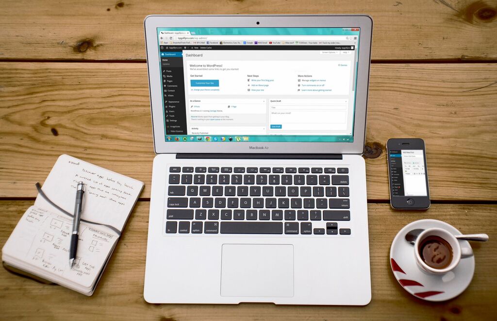 Ein silber-schwarzer Laptop auf dem ein Website-Dashboard zu sehen ist, steht auf einem Holztisch; daneben liegen ein offenes beschriebenes Notizbuch mit einem Kugelschreiber darauf, ein Smartphone sowie eine gefüllte Kaffeetasse.