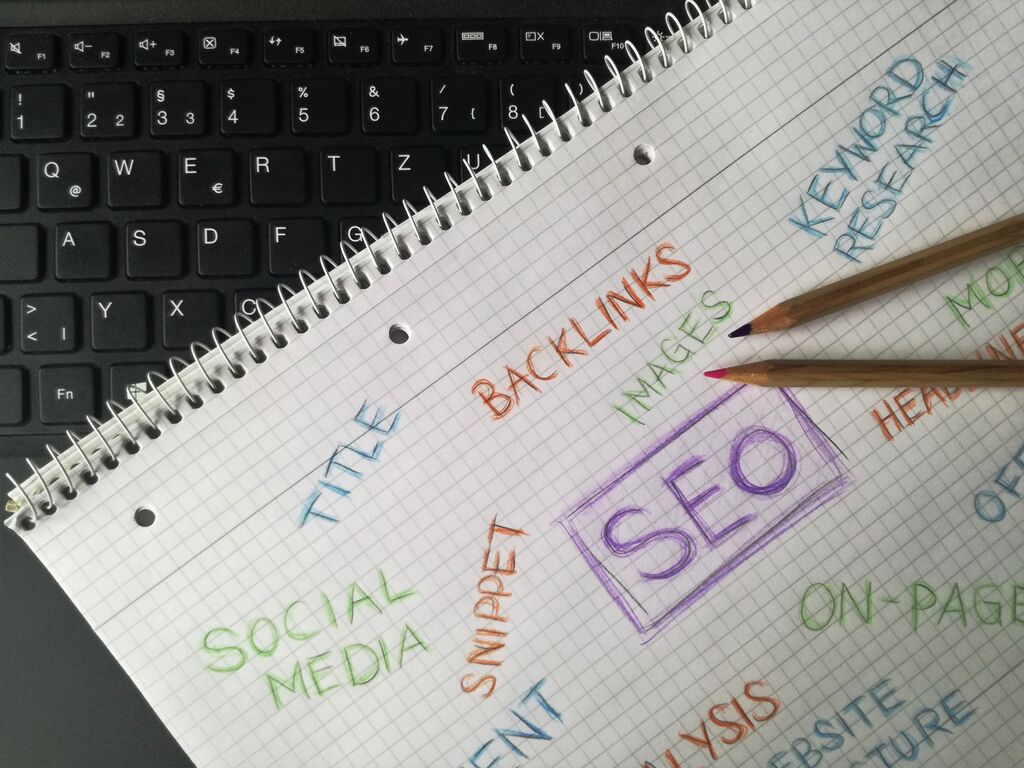 Ein Notizblock, auf dem einige bunte Keywords zum Thema SEO  um das zentrale Wort SEO herum geschrieben wurden, liegt auf einem Laptop.