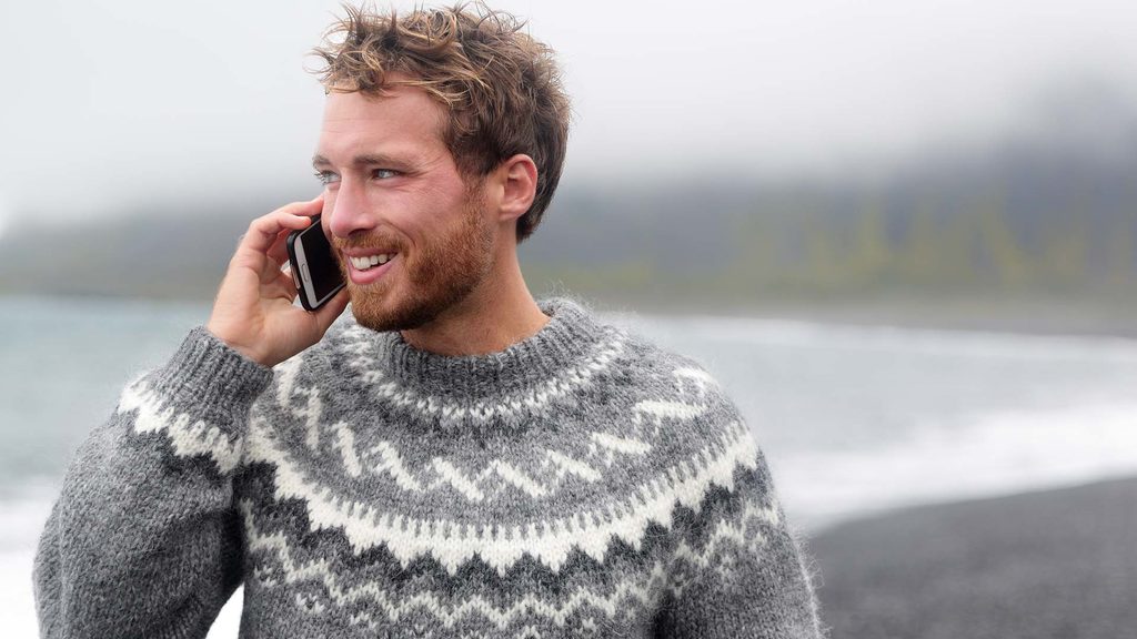 Mann som snakker i telefon med grå ullgennser og en strand i bakgrunnen med grått vær