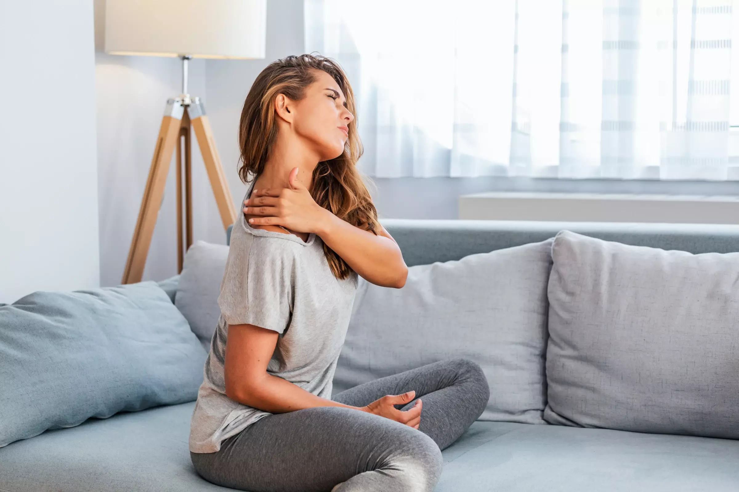 Frau sitzt mit Verspannungen und Schmerzen im Nacken auf einem Sofa