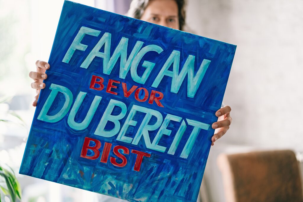 Werbeagentur für Startups Hamburg – Daniel Kaesmacher hält ein Bild mit einem Spruch in die Kamera: "Fang an, bevor Du bereit bist!"