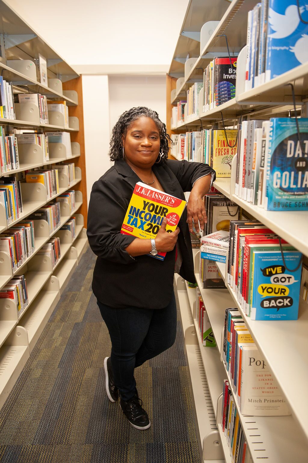 CEO Danielle "Dia" Winder holding a tax book while leaning on a bookshelf in the financial section of a library