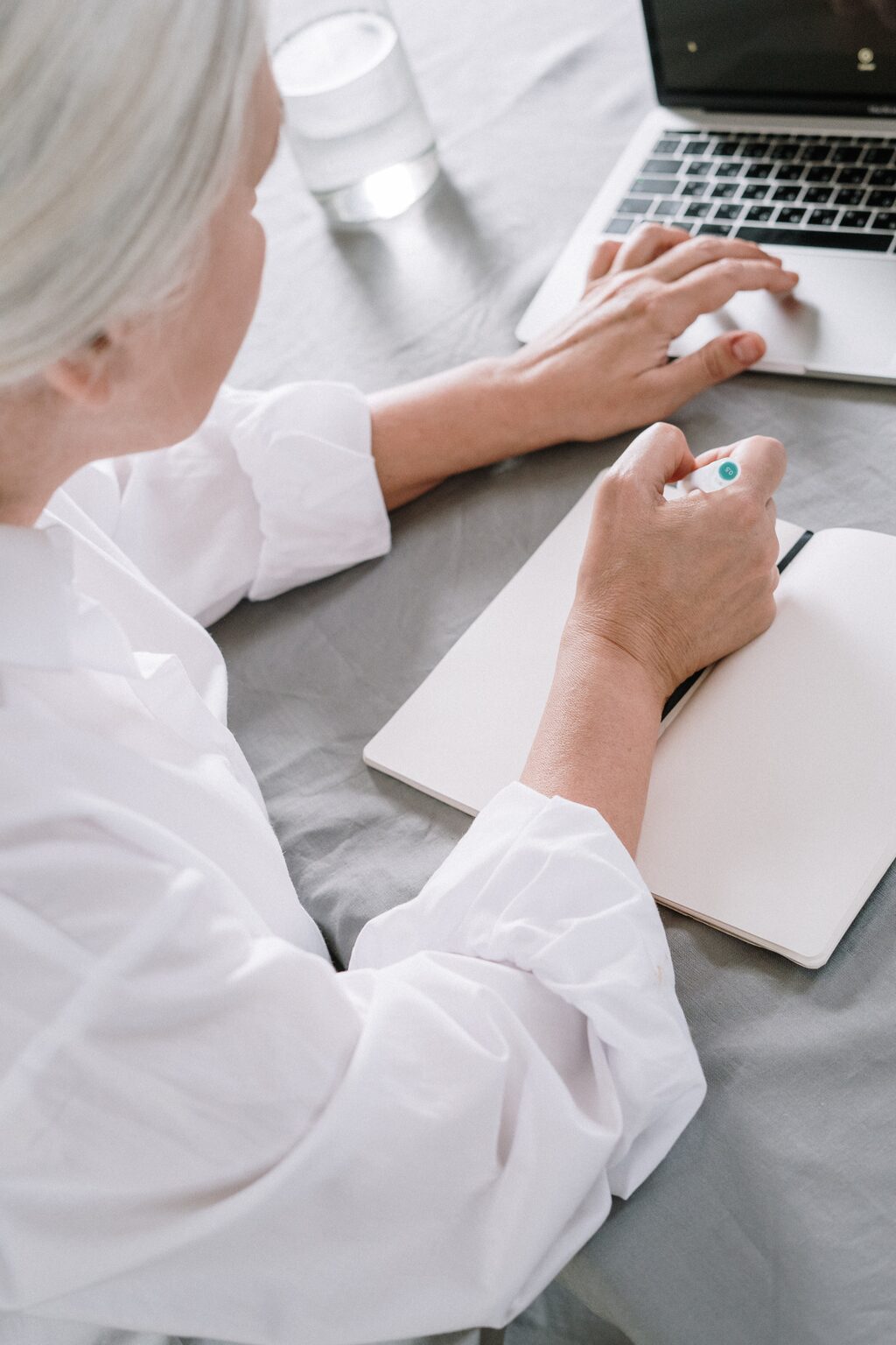 Elderly woman writing in notebook while on laptop