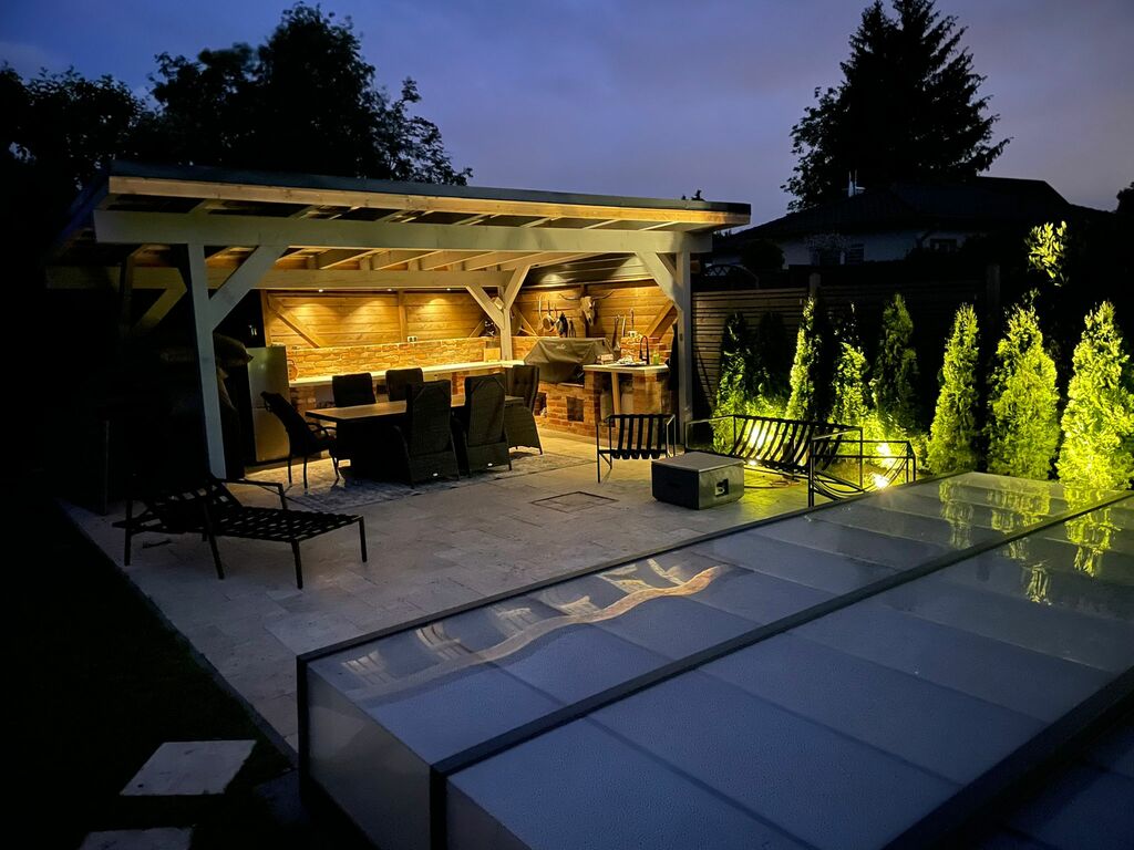 Abendaufnahme einer stilvollen Outdoor-Küche unter einem beleuchteten Holzpavillon, mit Esstisch und Lounge-Bereich auf einer Terrasse, umgeben von dezent beleuchteten Pflanzen