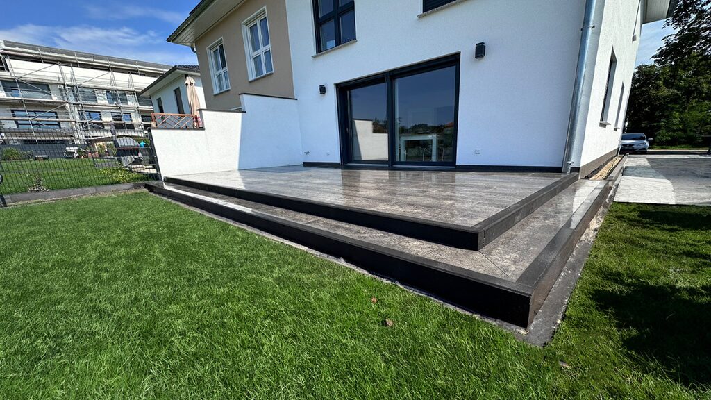 Großzügige, modern gestaltete Terrasse mit dunklem Rahmen und großformatigen Fliesen, mit mehreren Stufen zum Garten mit gepflegtem Rasen.