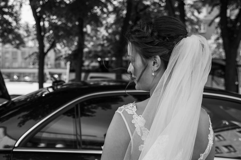 Black and white image of a woman in a white wedding dress standing with her back towards the camera. She is facing a black wedding car.