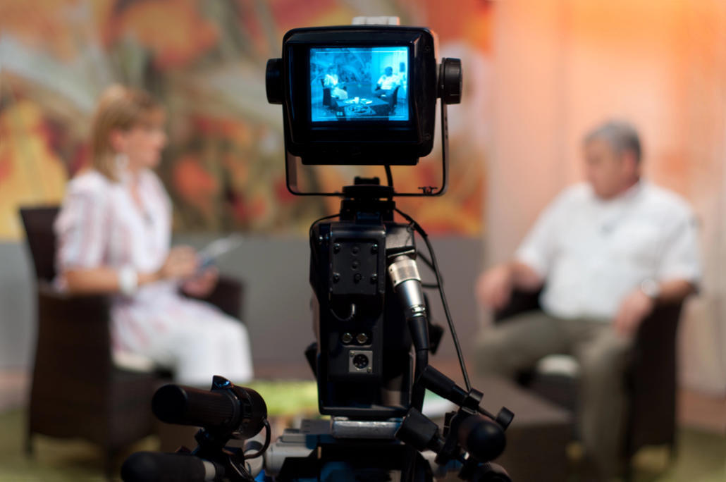 Studio video camera filming an interview; host and guest seated and blurred in the background, visible on the camera’s monitor.