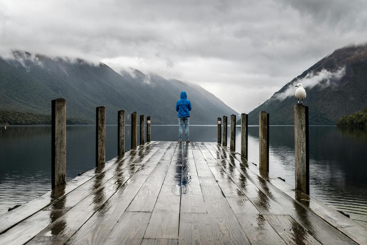 Person auf einem regennassen Steg mit Blick über einen See zu den Bergen