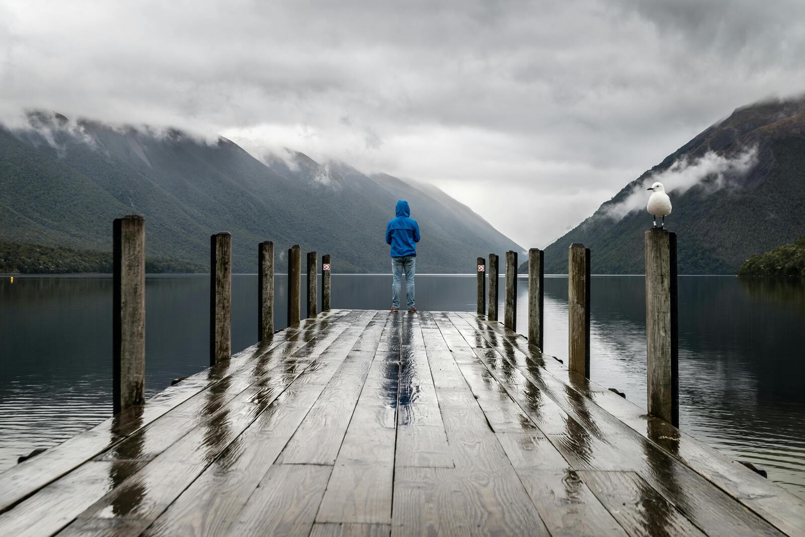 Person in blauer Jacke auf einem regennassen Steg, der Blick geht über einen stillen See zu den Bergen