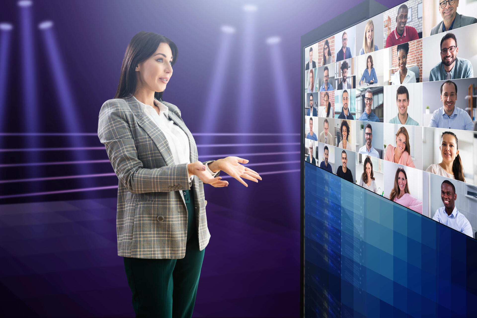 Presenter gestures toward a large monitor displaying a grid of remote meeting participants, set against a purple studio with spotlights.