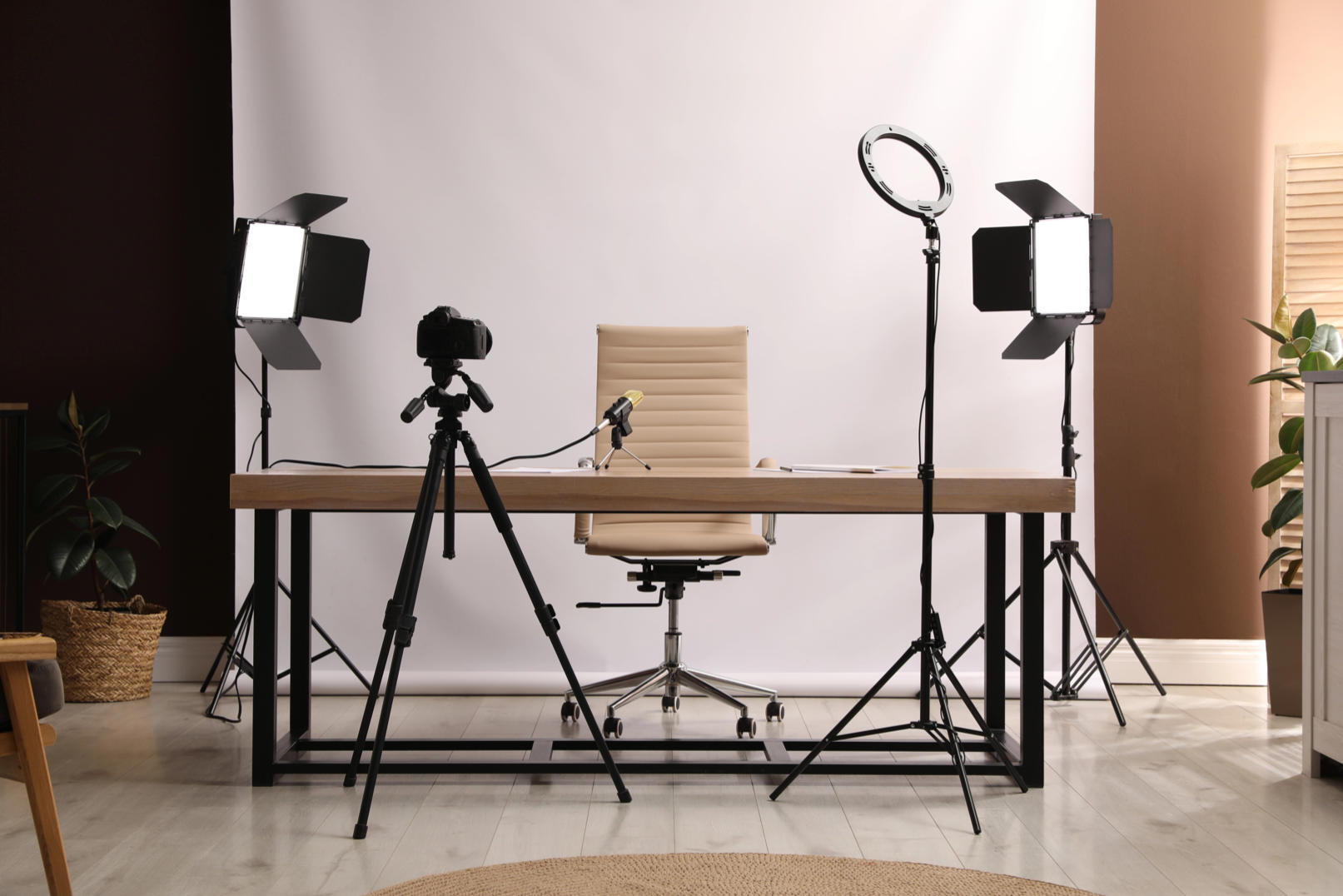 Home studio desk with beige chair, DSLR on tripod, microphone, ring light, and two softbox lights set against a white backdrop.