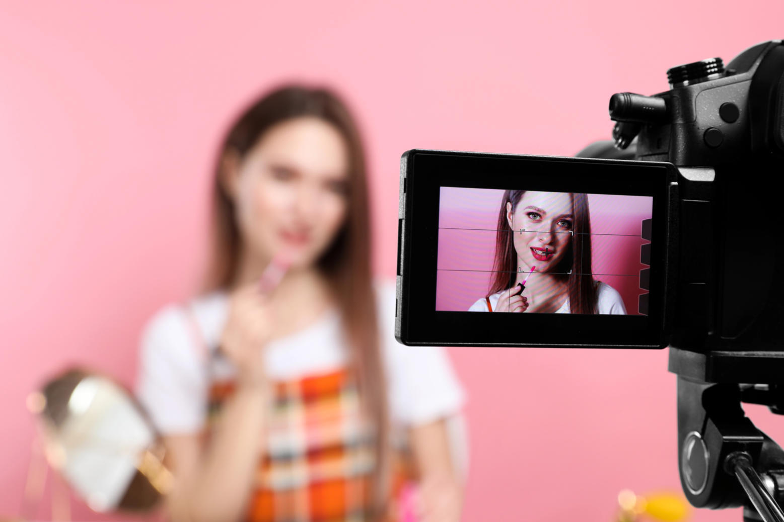 Camera screen in focus shows a woman applying pink lip gloss, while she appears blurred in the background against a pink backdrop.