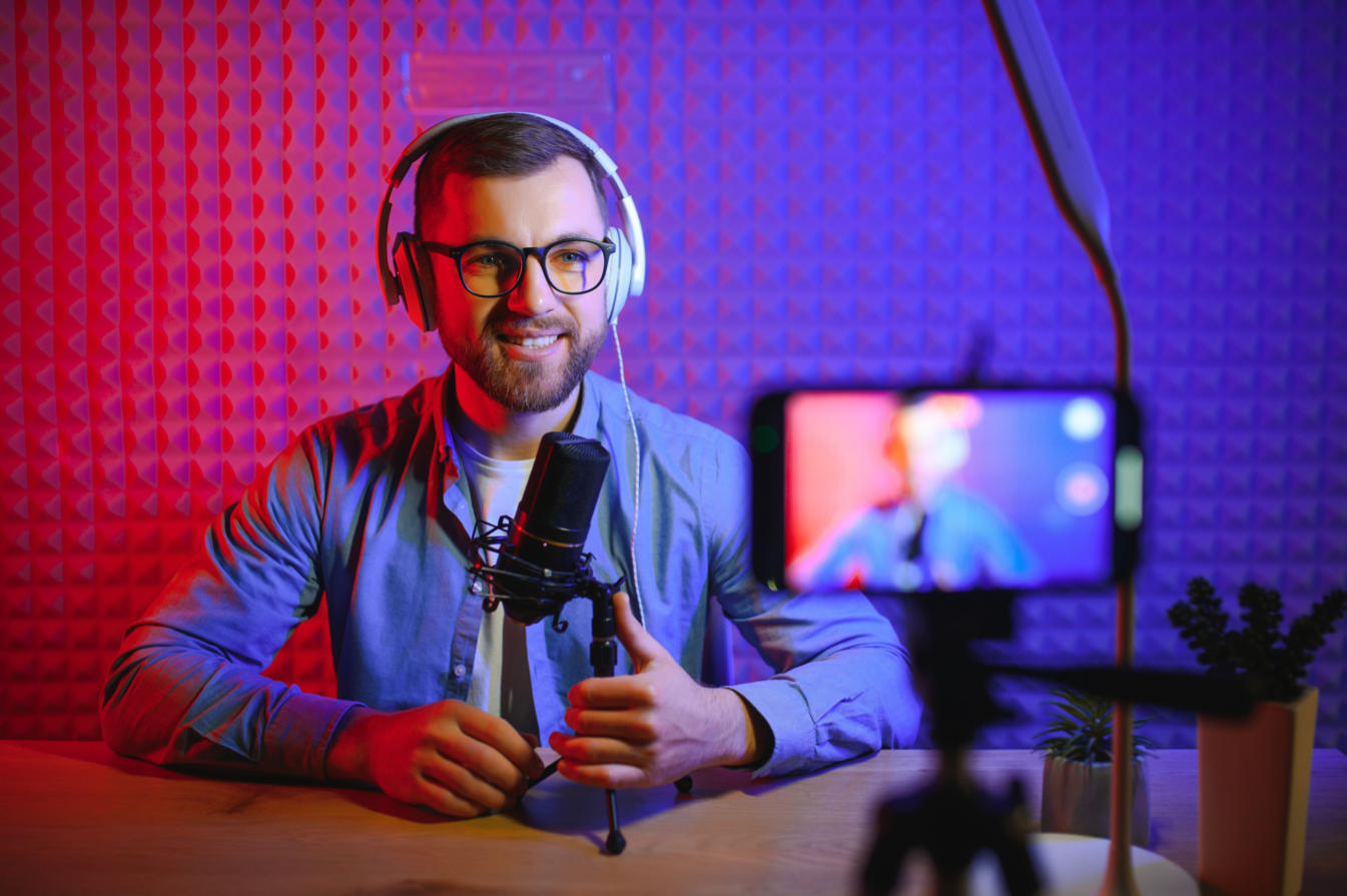 Smiling man with glasses and headphones talks into a desk mic; phone on a tripod films under blue and pink lighting.