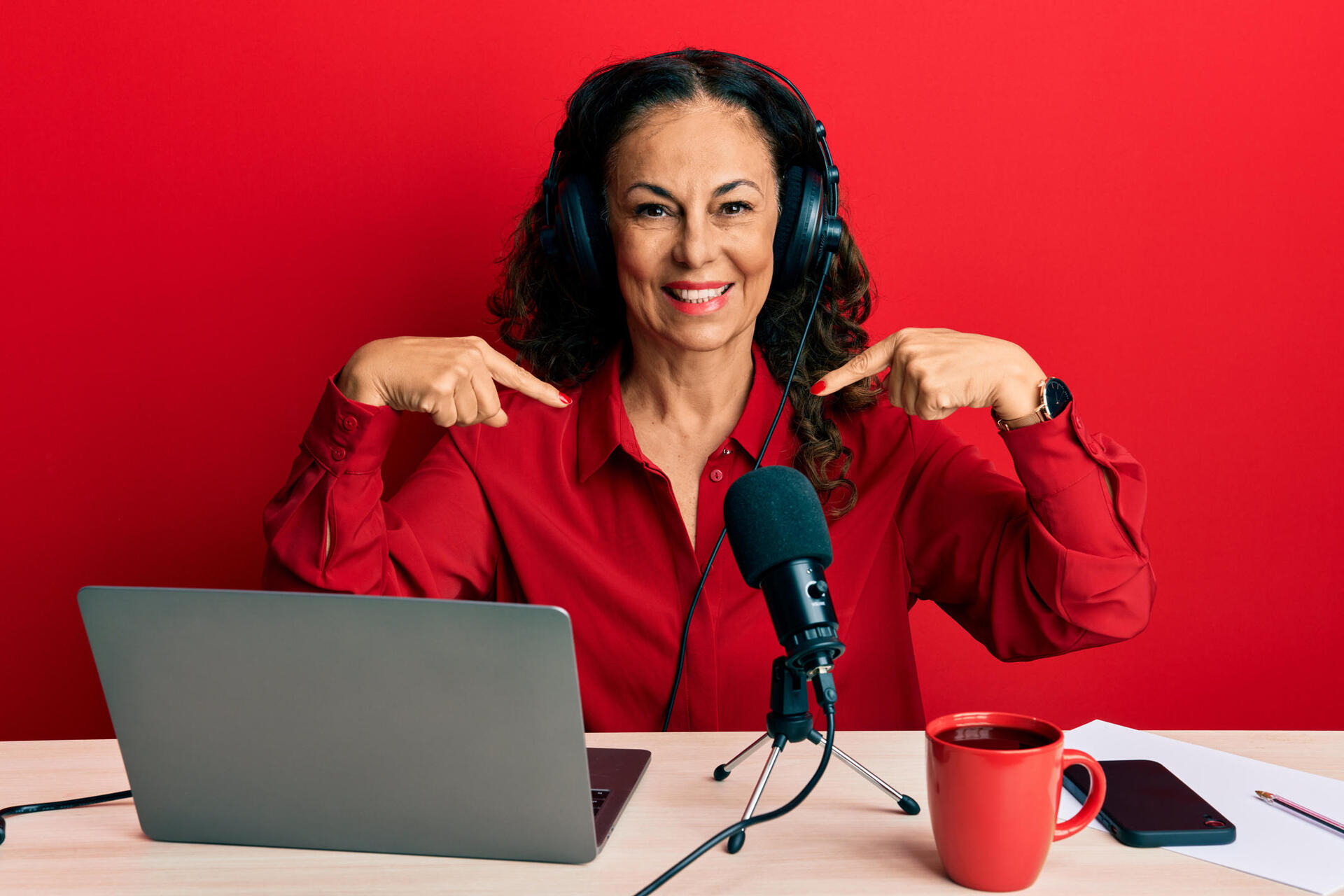 Smiling person in a red shirt and headphones points to themselves while recording at a desk with a laptop, microphone, phone, papers, and a red mug against a red background.