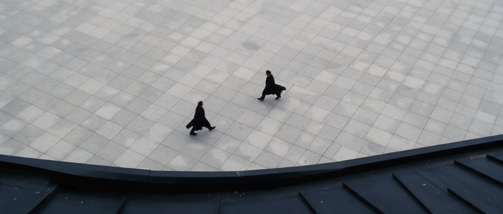 Zwei Figuren in schwarzen Mänteln kreuzen sich auf einem Steinplatz, von oben