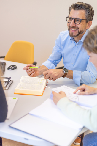 Personen sitzen an einem Tisch mit einem geöffneten Buch und Notizblöcken, eine Person hält einen grünen Stift.