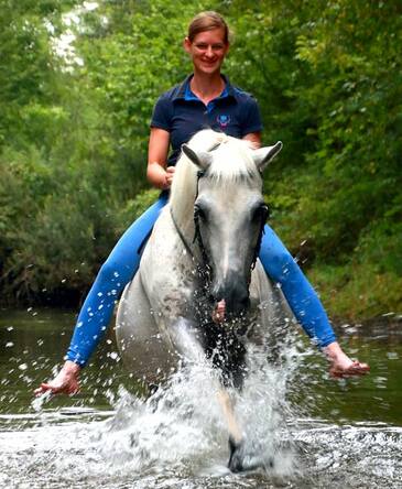 Steffi Hostettler auf einem Schimmel ohne Sattel durch einen Bach planschend
