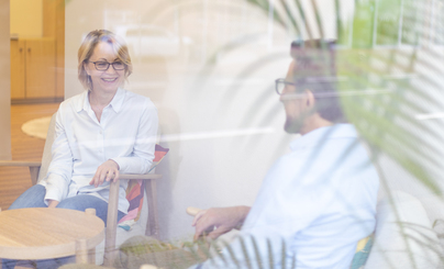 Zwei Personen sitzen sich an einem runden Holztisch in einem hellen Raum gegenüber, mit unscharfen grünen Pflanzen im Vordergrund.