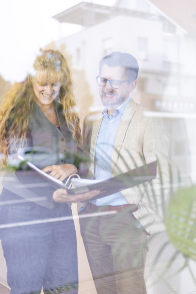 Dirk Kastner mit Mitarbeiterin stehend, halten gemeinsam einen Ordner in einem hellen Raum. 