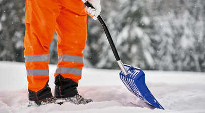 Arbeiter in reflektierender, orangefarbener Warnkleidung räumt Schnee mit einer blauen Schneeschaufel im winterlichen Außenbereich.