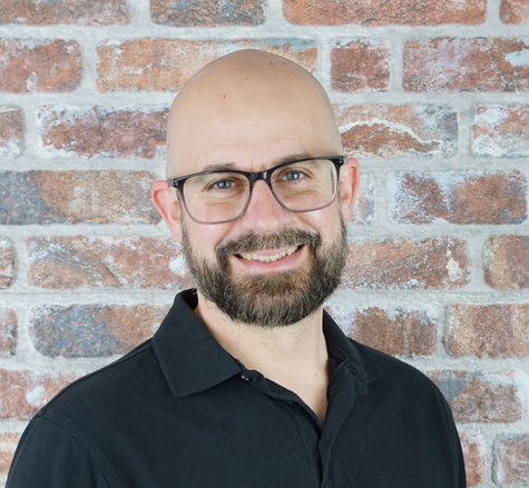 Porträtfoto von Christian Elia von Werbepixel. Der Mann mit Glatze, Bart und Brille trägt ein schwarzes Poloshirt und lächelt freundlich vor einer Backsteinwand.